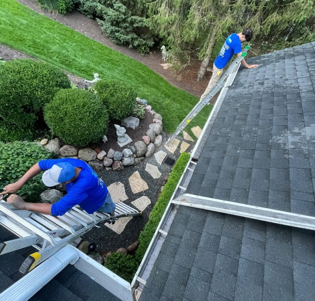 Two crew members cleaning out a home’s gutters from a ladder and roof edge